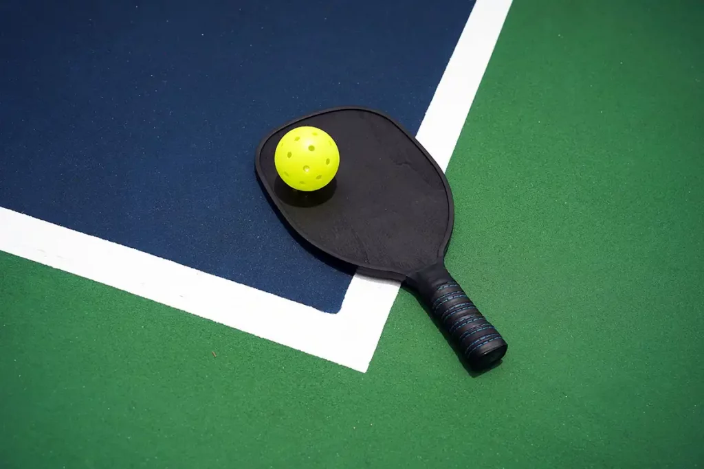 Pickleball paddle with a pickleball on top on the edge of a pickleball court